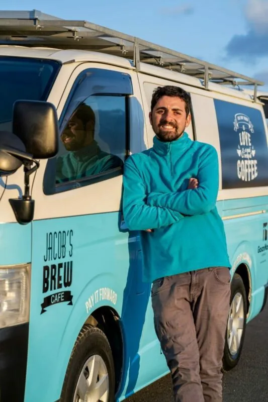 Jacob leaning against the turquoise Pay It Forward coffee van, solar panels on its roof and "Jacob's Brew Cafe — pay it forward" along the side